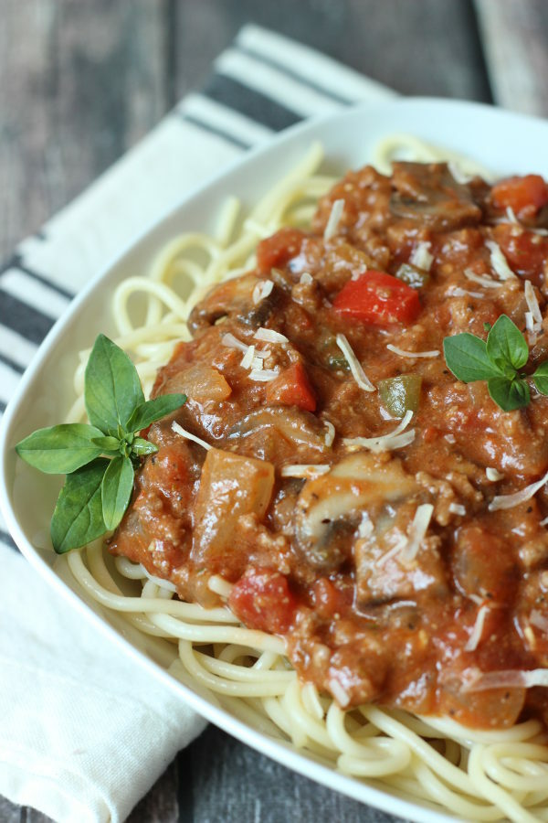 A big square bowl is full of spaghetti noodles and topped with a meaty, chunky spaghetti sauce. There are some basil leaves as garnish and a cloth napkin on the table ready to sop up some tomato sauce.