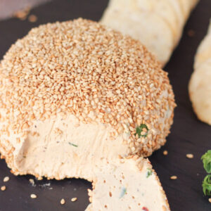 A lovely Asian cheese ball has been rolled in toasted sesame seeds and set on a black stone serving board. You can see one slice out of the ball and gluten free rice crackers ready for serving.