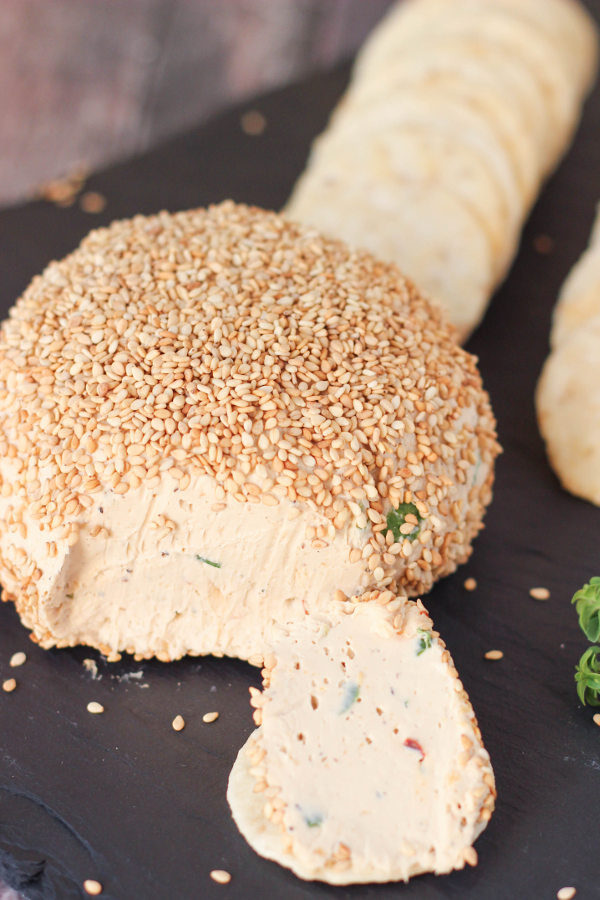 A lovely Asian cheese ball has been rolled in toasted sesame seeds and set on a black stone serving board. You can see one slice out of the ball and gluten free rice crackers ready for serving.