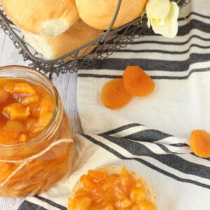 A fresh jar of dried apricot jam sits next to an English muffin slathered with jam. In the background a wire basked full of rolls and roses waits for a hungry guest.