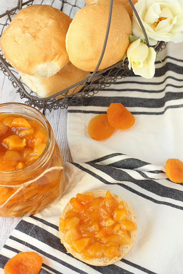 A fresh jar of dried apricot jam sits next to an English muffin slathered with jam. In the background a wire basked full of rolls and roses waits for a hungry guest.