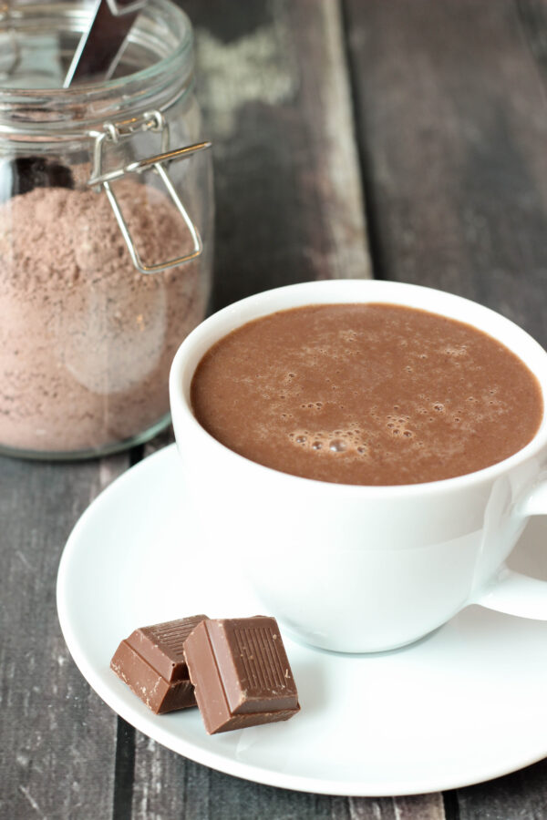 A large white cup of hot chocolate sits on a plate with a few squares of chocolate. Behind you see a glass jar of triple chocolate hot cocoa mix.