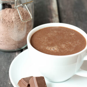 A large white cup of hot chocolate sits on a plate with a few squares of chocolate. Behind you see a glass jar of triple chocolate hot cocoa mix.