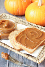 Slices of toast on a white platter are slathered with spicy homemade pumpkin butter. You can see pie pumpkins in the background and a mason jar with additional pumpkin butter.