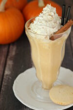 A clear ice cream soda fountain glass is full of a boozy pumpkin pie milkshake and topped with whipped cream and a stick of cinnamon. In the background you see pumpkins.