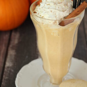 A clear ice cream soda fountain glass is full of a boozy pumpkin pie milkshake and topped with whipped cream and a stick of cinnamon. In the background you see pumpkins.