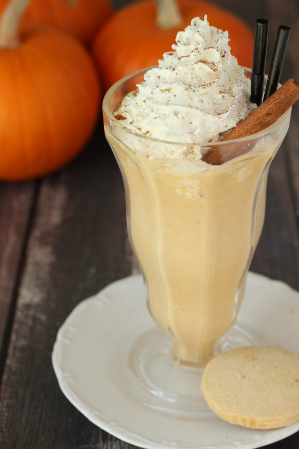 A clear ice cream soda fountain glass is full of a boozy pumpkin pie milkshake and topped with whipped cream and a stick of cinnamon. In the background you see pumpkins.