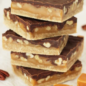 Five big turtle bars are stacked up on a table. You can see a big layer of chocolate, a thick layer of caramel full of pecans and then a layer of shortbread cookie on the bottom.