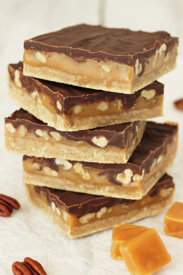 Five big turtle bars are stacked up on a table. You can see a big layer of chocolate, a thick layer of caramel full of pecans and then a layer of shortbread cookie on the bottom.