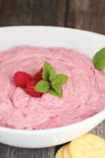 A white serving bowl is full of cream cheese cranberry raspberry dip. Red raspberries and mint sprigs garnish the top of this Thanksgiving appitizer.