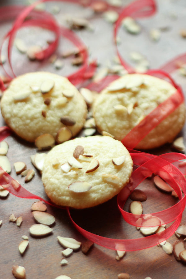 A wooden table has three almond cookies in the middle of it. There are slivered almonds scattered around and some gauzy red ribbon swirls.