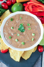 A white bowl of creamy black bean dip is garnished with green onions. It is surrounded by red peppers, cherry tomatoes, chips and wedges of lime.