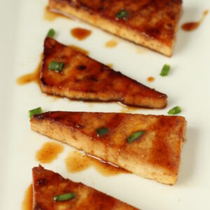 fried teriyaki tofu triangles are lined up on a white platter. There is a sprinkling of green onions garnishing it.