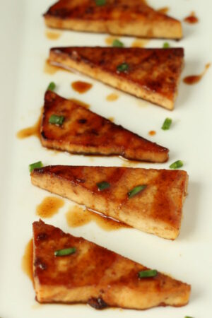 fried teriyaki tofu triangles are lined up on a white platter. There is a sprinkling of green onions garnishing it.