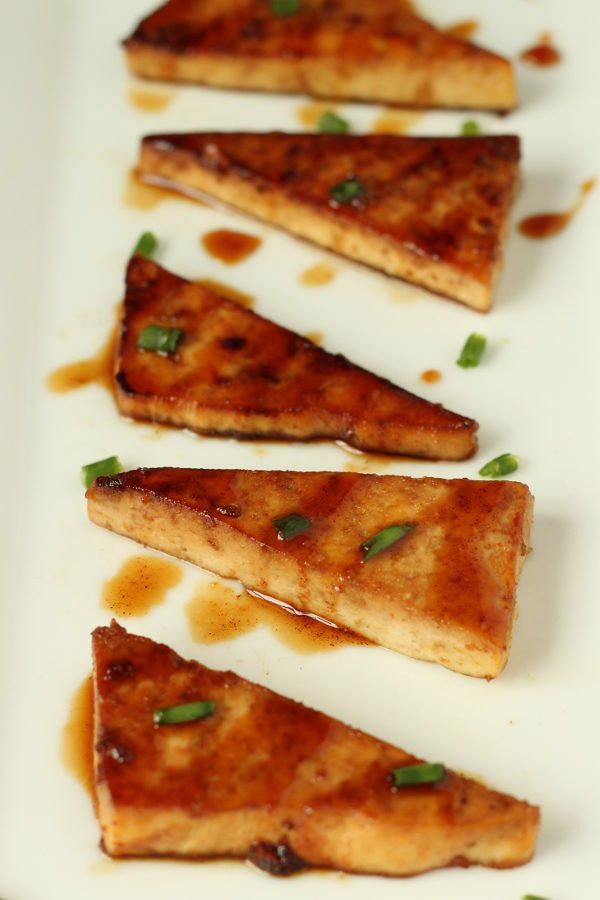 fried teriyaki tofu triangles are lined up on a white platter. There is a sprinkling of green onions garnishing it.