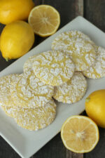 A square white platter is piled up with pretty lemon snowflake cookies. The cookies are yellow with a powdered sugar snowflake pattern on them. The platter is surrounded by lemons sitting on a wooden table.