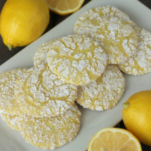 A square white platter is piled up with pretty lemon snowflake cookies. The cookies are yellow with a powdered sugar snowflake pattern on them. The platter is surrounded by lemons sitting on a wooden table.