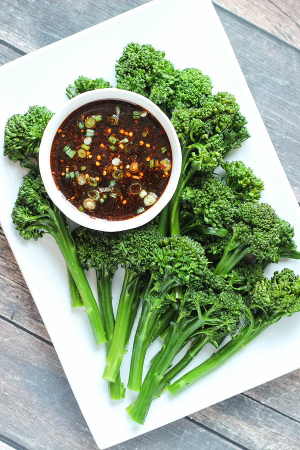 A white platter is full of lovely broccoli spears, perfectly steamed and brilliant green. At the top a bowl of korean bbq sauce waits for the broccoli to be dipped. In the sauce you can see sliced green onion, red pepper, and other spices in the soy sauce base.