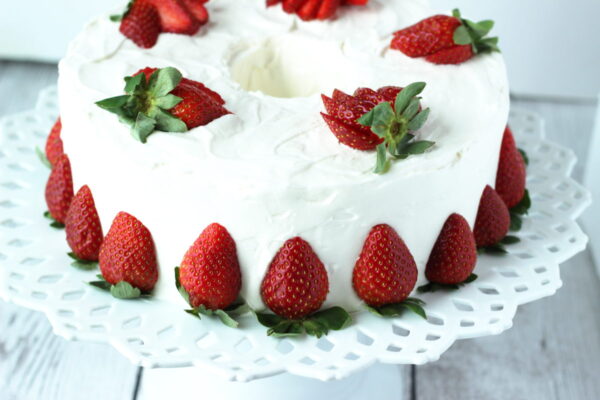 A pound cake is frosted with snow white whipped cream frosting. The cake is siting on an elegant white ceramic cake plate. There are cut strawberries around the base of the cake and sliced strawberries fanned out on top.