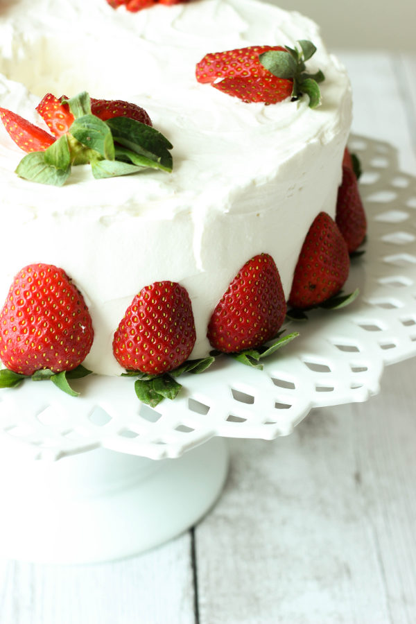 A stunning cake sits on decorative white cake stand. The cake is simple frosted with whipped cream frosting and decorated with fresh strawberries sliced or cut in half.
