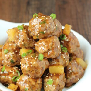 A white plate is stacked high with delicious teriyaki meatballs. There are chunks of pineapple in the sauce and some green onions.