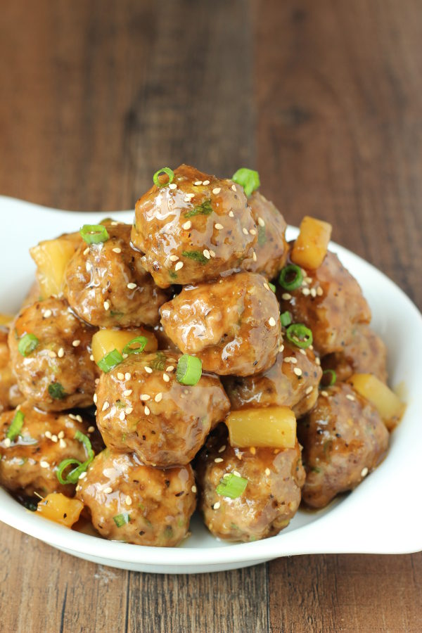 A white plate is stacked high with delicious teriyaki meatballs. There are chunks of pineapple in the sauce and some green onions.
