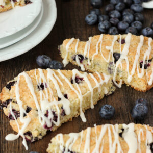Three fresh blueberry scones sit on a table next to a stack of white plates. A pile of blueberries is spilling out of a dish among the scones.