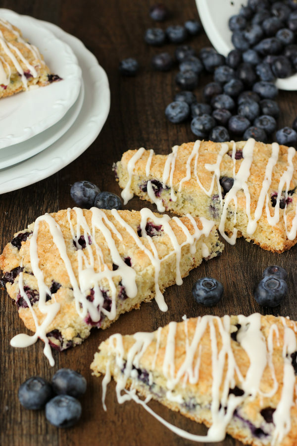 Three fresh blueberry scones sit on a table next to a stack of white plates. A pile of blueberries is spilling out of a dish among the scones.
