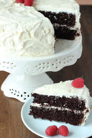 A chocolate cake sits on a white cake stand. The cake is frosted with vanilla buttercream.