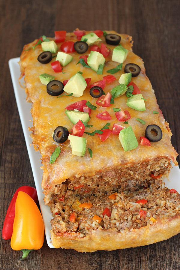 A big loaf of Mexican meatloaf rests on a white platter. It is garnished with olives, tomatoes and avocados.