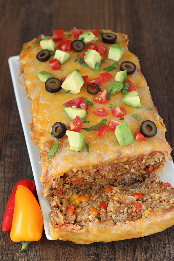 A big Mexican meatloaf sits on a white platter. The first slice has been cut off and you can see a thick layer of cheese on top of a rich meatloaf studded with chunks of bell peppers and plenty of rice.