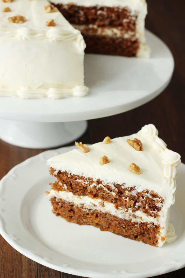 A carrot cake sits on a white cake pedestal. The cake is frosted with fluffy cream cheese frosting and decorated with a few walnuts. A wedge of cake sits next to the stand ready to eat.