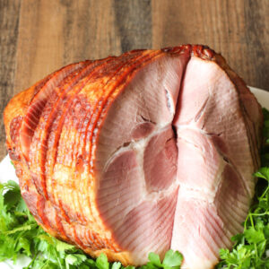 A large spiral ham is sitting on a bed of greenery and white platter waiting to be served for the holidays.