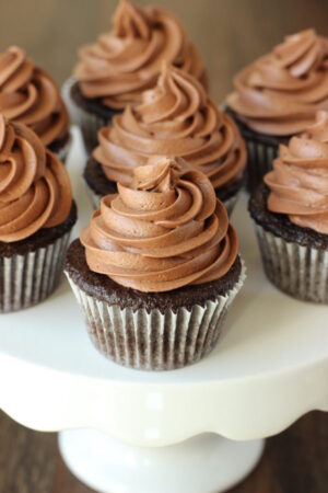 Chocolate cupcakes topped with giant swirls of chocolate cheesecake frosting sit on a white cake stand waiting to be enjoyed.