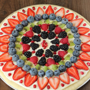A fruit pizza made from a no chill sugar cookie crust, cream cheese frosting, and fruit. It is decorated with pomegranates, blackberries, raspberries, kiwi, blueberries, and sliced strawberries.