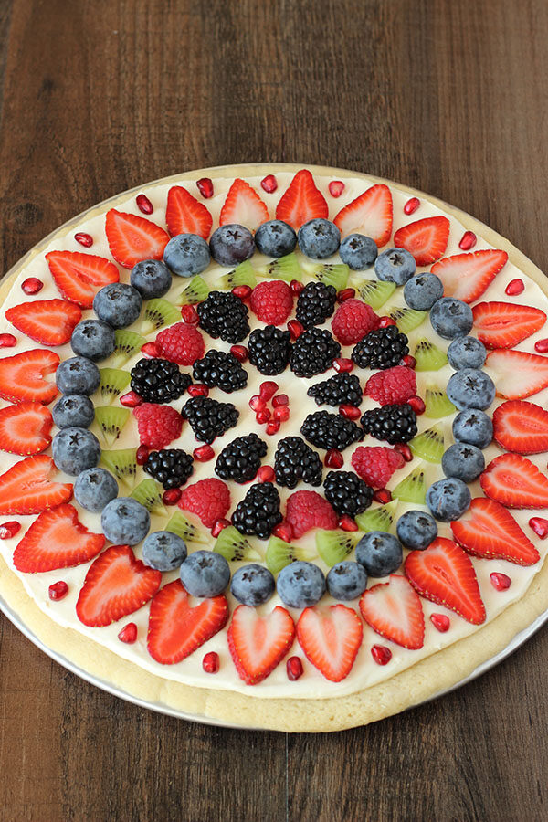A fruit pizza made from a no chill sugar cookie crust, cream cheese frosting, and fruit. It is decorated with pomegranates, blackberries, raspberries, kiwi, blueberries, and sliced strawberries. 