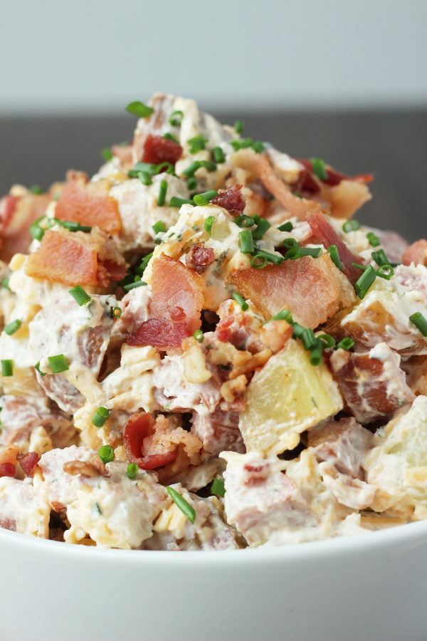 A big white bowl of loaded potato salad. The salad is full of chunks of sausage and pieces of bacon and covered in chives.