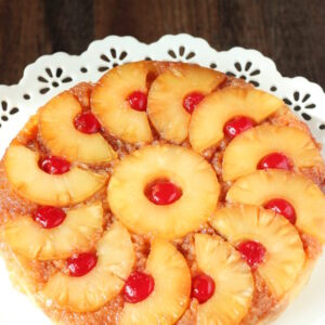 A beautiful pineapple upside down cake is displayed on a white cake stand. The pineapple is cut into half circles and arranged like a pinwheel around a center ring of pineapple. Cherries sit inside each ring or half ring.