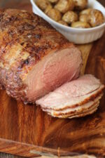 A roasted leg of lamb with lovely brown crust is cut into slices on a cutting board. Behind you see a dish of roasted new potatoes.