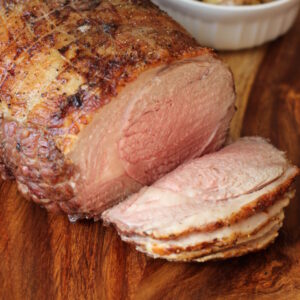 A roasted leg of lamb with lovely brown crust is cut into slices on a cutting board. Behind you see a dish of roasted new potatoes.