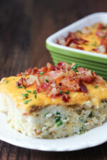 A big slice of loaded twice baked potato casserole sits on a white plate. It is covered in cheese, bacon and chives.