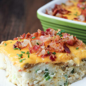A big slice of loaded twice baked potato casserole sits on a white plate. It is covered in cheese, bacon and chives.