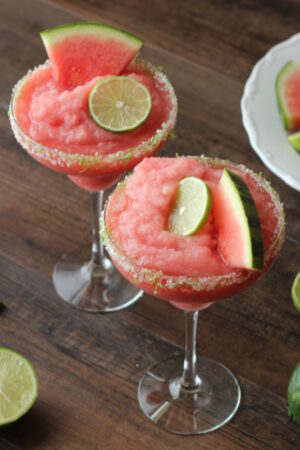 Two frosty glasses of frozen watermelon margaritas garnished with wedges of watermelon and slices of lime.
