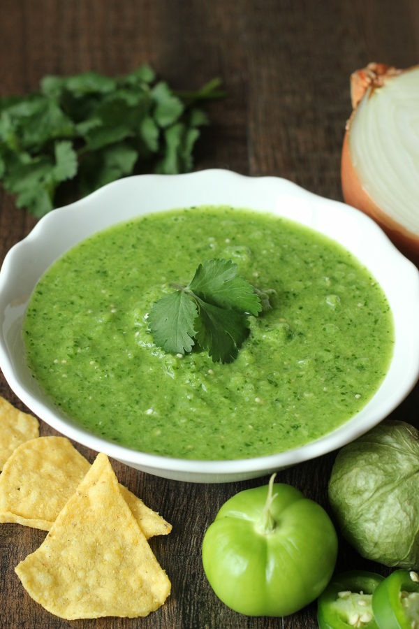 A beautiful bowl of bright green salsa verde sauce sits on the table. The sauce is garnished with a cilantro leaf and surrounded by tomatillos, chips, cilantro, and a cut onion.