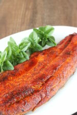A white platter with a salmon filet on it. The salmon is seasoned with a brown sugar glaze and garnished with some basil.