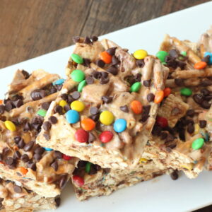 A white platter is sitting on a wooden table. The platter is full of marshmallow treat bar squares. You can see rice chex and pretzels in the bars. The tops are sprinkled with mini m and ms and mini chocolate chips.