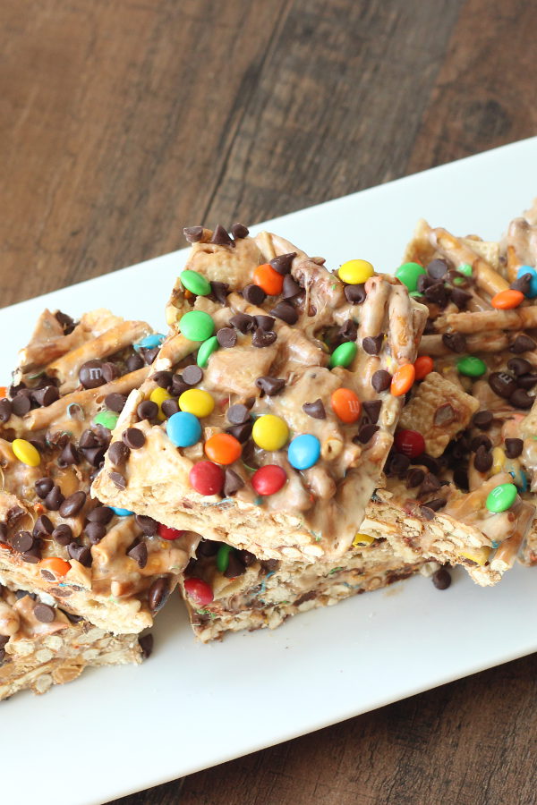 A white platter is sitting on a wooden table. The platter is full of marshmallow treat bar squares. You can see rice chex and pretzels in the bars. The tops are sprinkled with mini m and ms and mini chocolate chips.