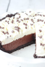 A beautiful chocolate cream pie is in a white pie plate. There is one slice removed so you can see the chocolate cookie crust, the rich chocolate filling, and swirls of whipped cream on top. There are mini chocolate chips scattered over the pie.
