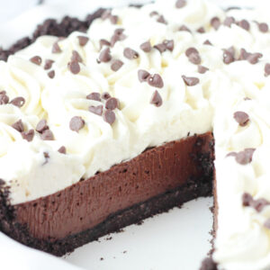 A beautiful chocolate cream pie is in a white pie plate. There is one slice removed so you can see the chocolate cookie crust, the rich chocolate filling, and swirls of whipped cream on top. There are mini chocolate chips scattered over the pie.