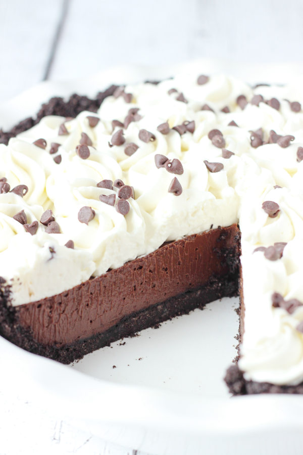 A beautiful chocolate cream pie is in a white pie plate. There is one slice removed so you can see the chocolate cookie crust, the rich chocolate filling, and swirls of whipped cream on top. There are mini chocolate chips scattered over the pie.